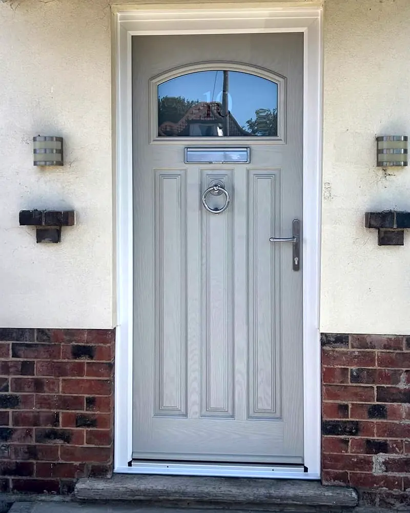 A pale grey composite front door with an arched glass panel, a ring knocker and a chrome lever handle, in a porch.