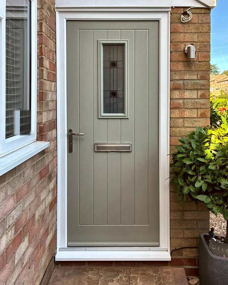 A sage green cottage-style composite front door with a slim leaded glass panel, beside a bay tree in a pot.