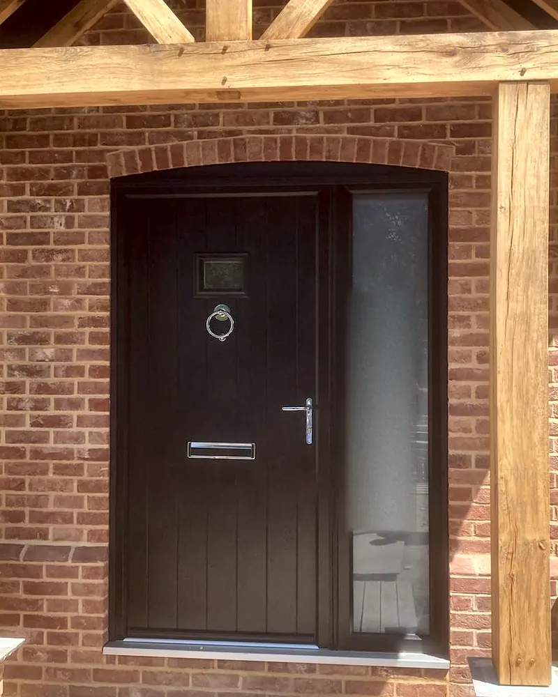 A dark brown cottage-style composite front door with a small glass panel and a glazed side panel, under an oak-framed porch.