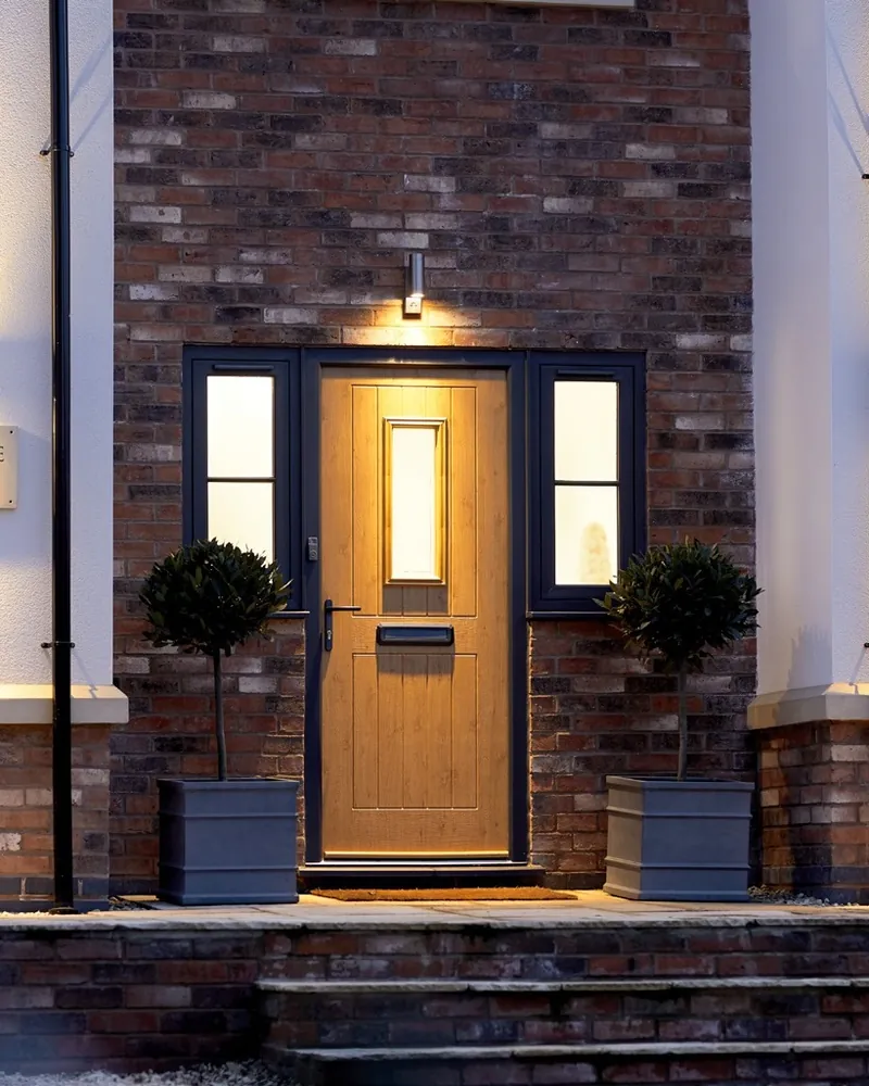 An oak-effect Endurance front door between two windows, lit by wall lights at dusk.