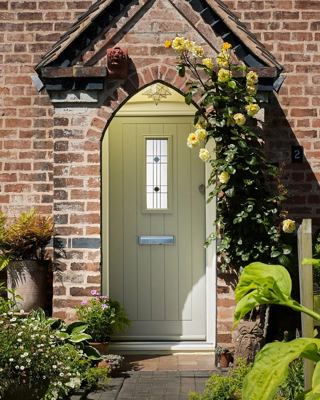 A sage green Endurance front door under a brick arch, with climbing roses and a cottage garden.