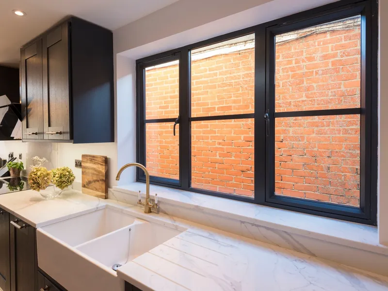 Black aluminium flush casement windows over a white butler sink and a marble worktop.