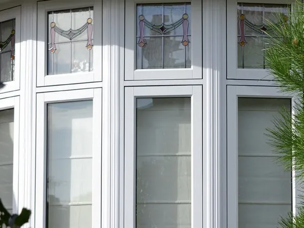A white uPVC bay window with leaded, stained-glass top lights, beside a pine tree.