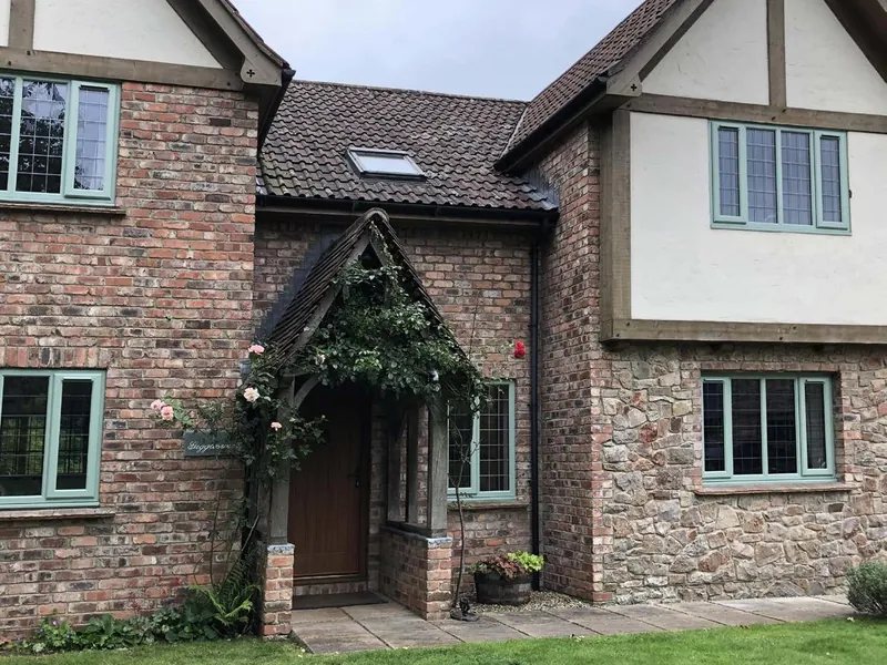 A brick, stone and timber-framed house with Chartwell Green casement windows and an oak porch with roses.