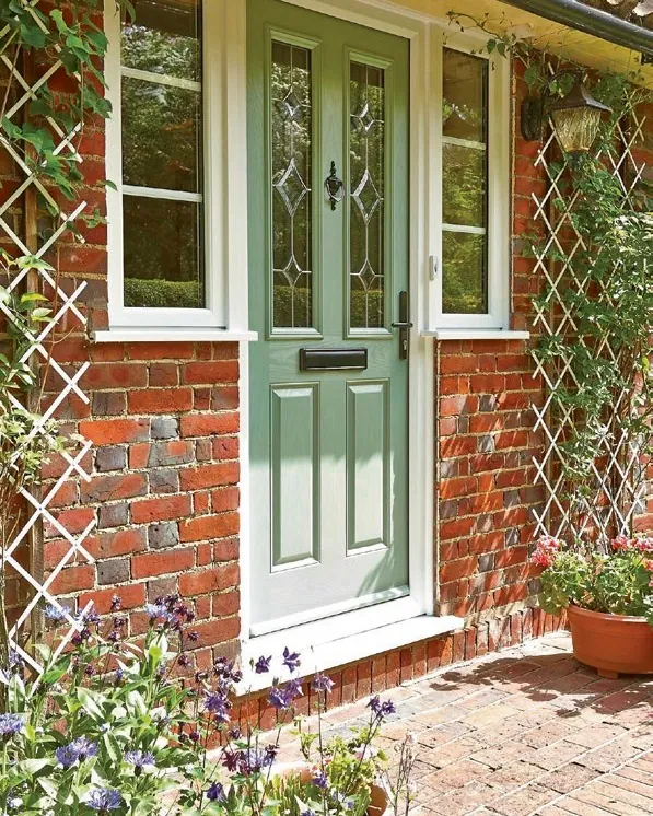 A sage green front door with decorative glass, between white windows and a garden trellis.