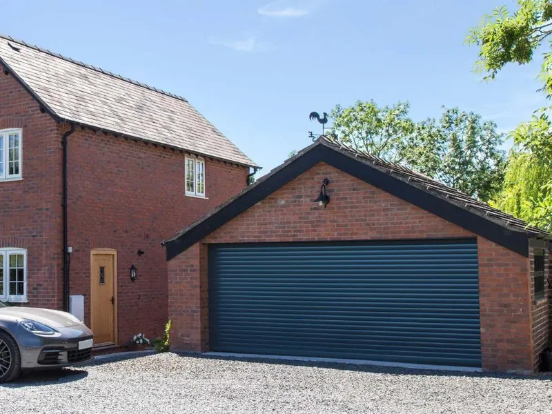 A brick house beside a detached double garage, closed by a wide dark grey roller door, with a car on the gravel drive.