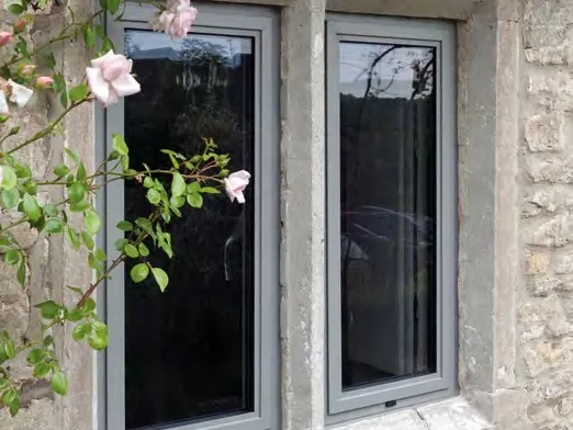 Two slim grey aluminium casement windows set in a stone wall, with a pink climbing rose.