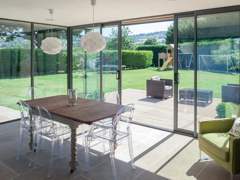 Slim grey aluminium sliding doors across a dining room, open onto decking and a lawn.