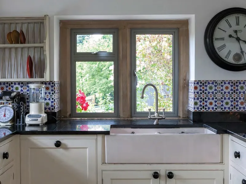 A pair of aluminium casement windows over a white butler sink, with patterned tiles and a wall clock.