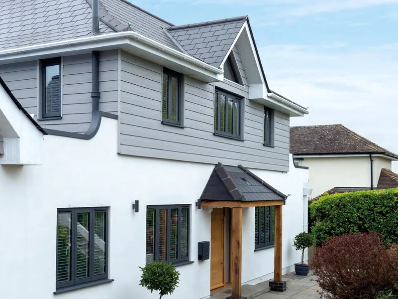 A white rendered house with dark grey aluminium windows and an oak front door under a porch.