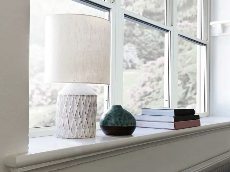 A white window with a lamp and books on its deep sill, looking out to a garden.