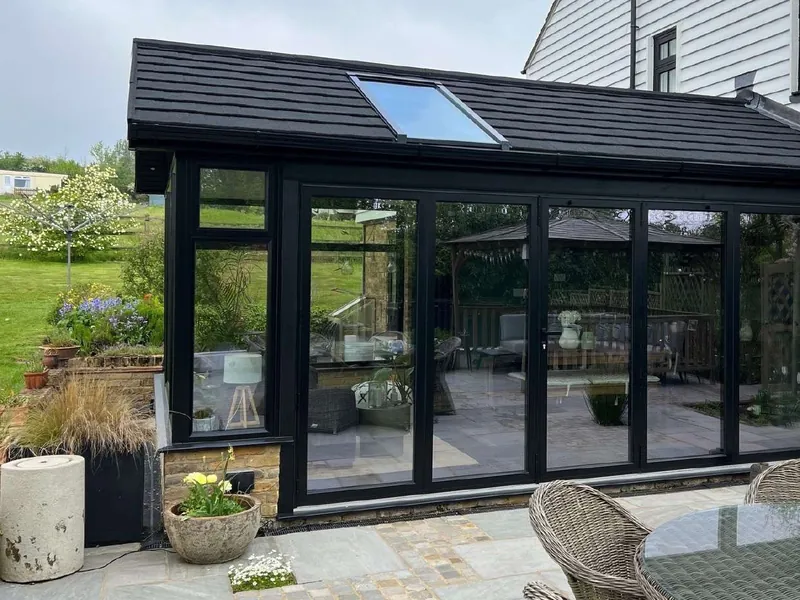 A black-framed room with a black tiled roof and a roof window, glazed across the front, beside a white weatherboarded house, with a patio in front.