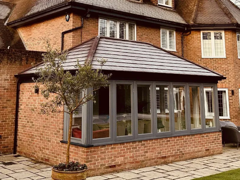 A grey-framed conservatory with a red-brick base and a hipped tiled roof, on a large brick house, with an olive tree in a pot.