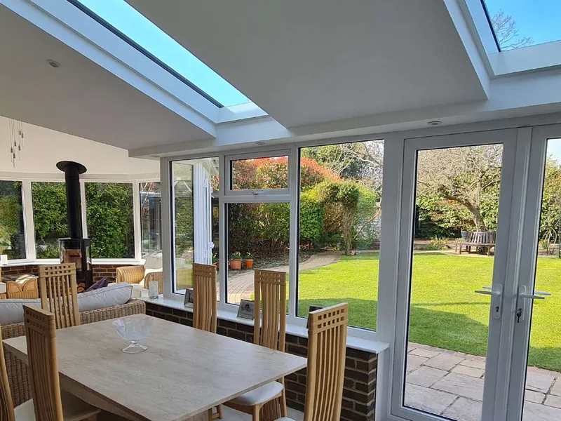 Inside a conservatory under a tiled roof with glass panels: a dining table, French doors open to the lawn and a log burner in the faceted end.
