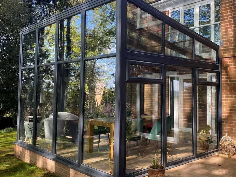 A dark-framed, fully glazed lean-to extension against a brick house, in a leafy garden.
