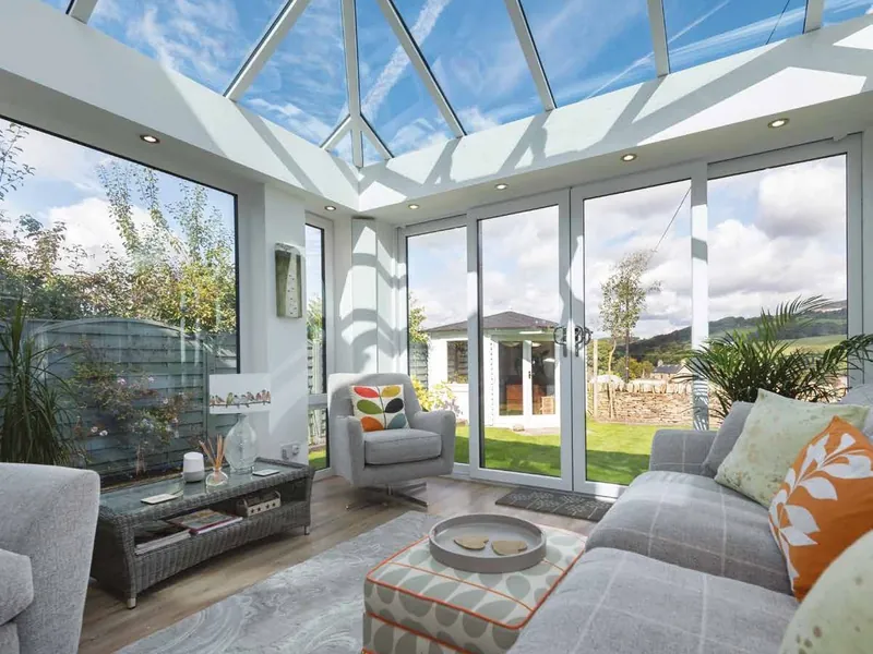 A bright living room under a glazed conservatory roof, with sofas, a rug and doors open to the garden.