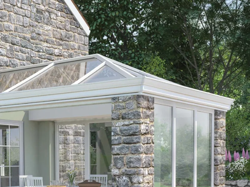A white orangery with stone corner piers, wide glazing and a roof lantern, beside a stone house.