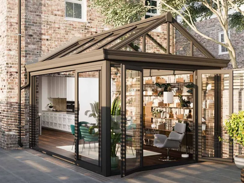 A conservatory in bronze-coloured frames with a glass roof and a glazed gable front, its doors folded open to a patio, against a brick house.