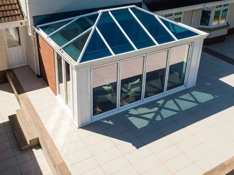 A white conservatory with a hipped, blue-tinted glass roof, seen from above beside a patio.