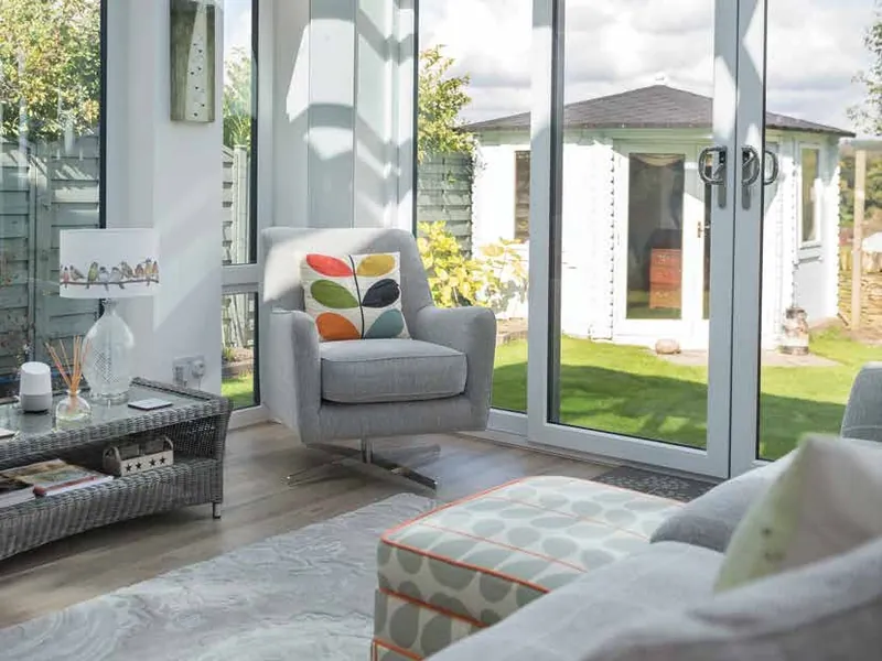 A bright white conservatory with a grey sofa, its doors open to the garden.
